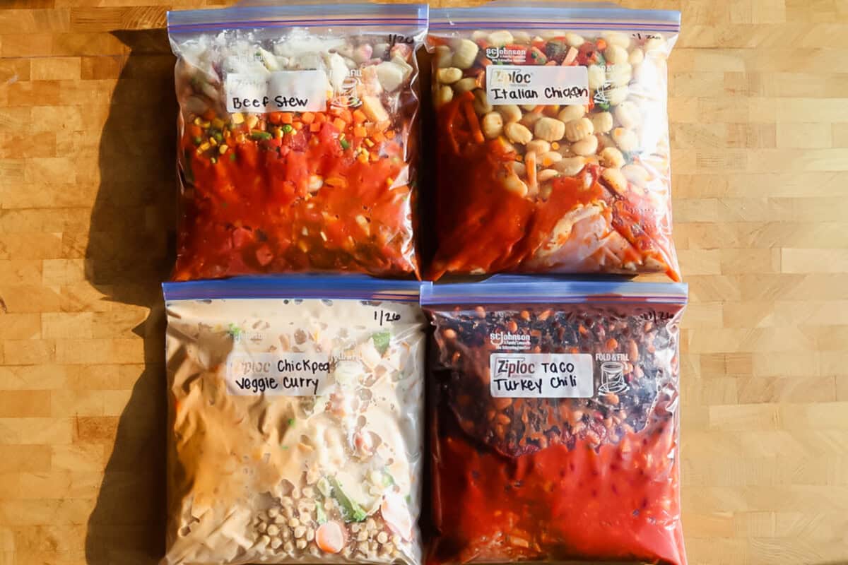 Four labeled freezer bags filled with homemade meals—beef stew, Italian chicken, chickpea veggie curry, and taco turkey chili—arranged on a wooden surface.
