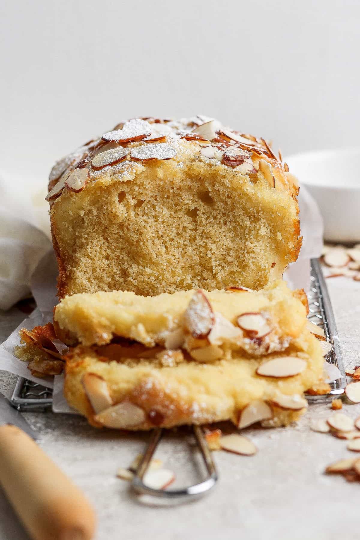 A loaf of almond cake with two slices cut, topped with sliced almonds and powdered sugar, rests on a wire rack beside a knife—evoking the cozy charm of cottage cheese pull apart bread.
