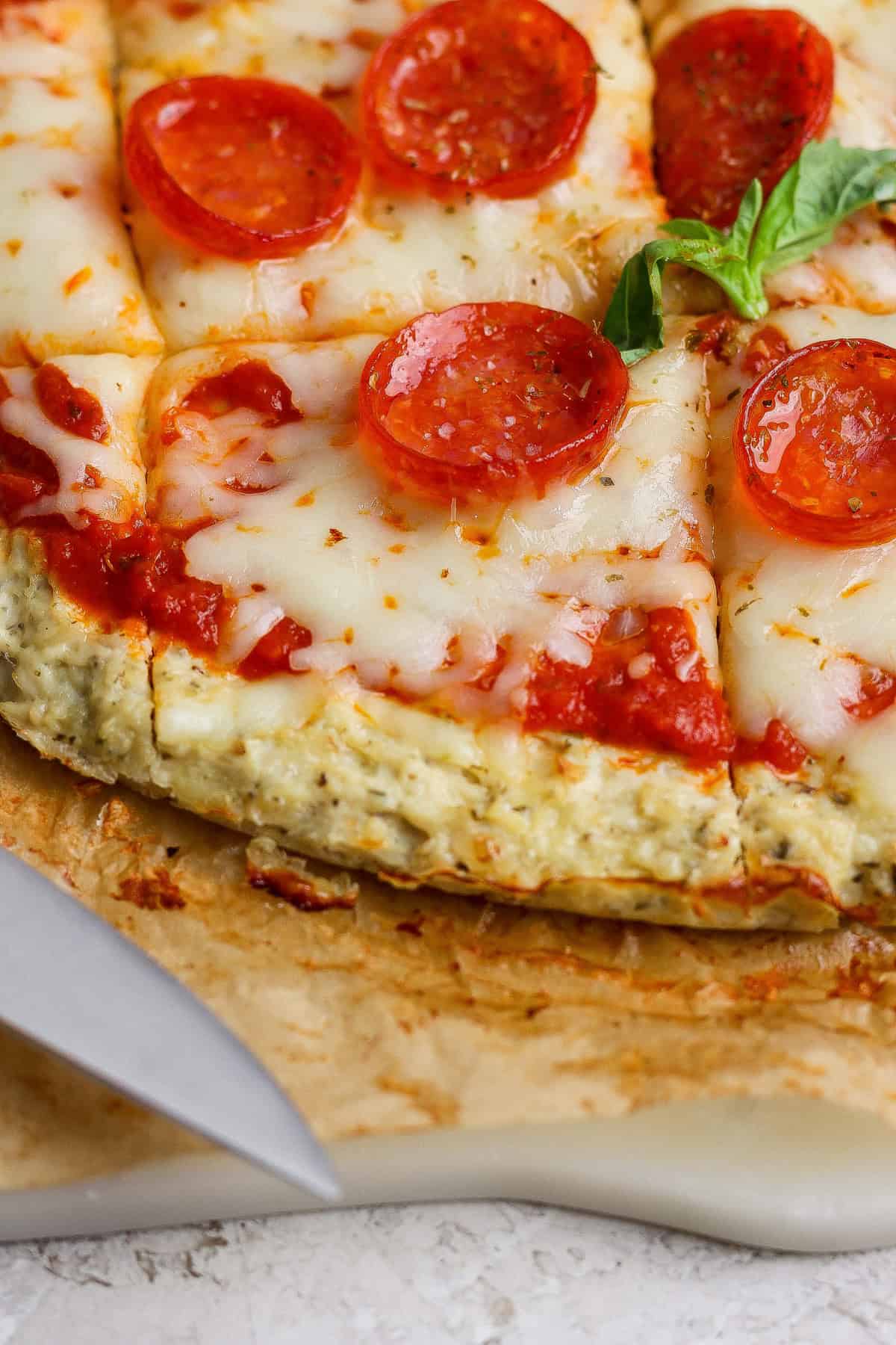Close-up of a sliced chicken crust pizza with melted cheese and tomato sauce on a cutting board, garnished with a sprig of basil. A knife rests nearby.