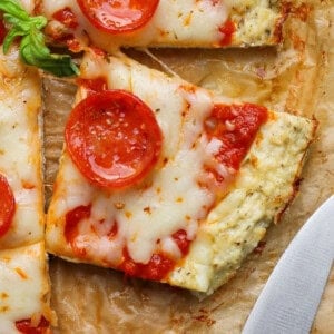A square slice of pepperoni chicken crust pizza with melted cheese and tomato sauce on parchment paper, next to a knife.