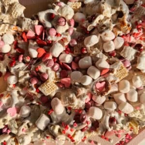 A close-up of a valentines day snack mix with cereal squares, mini marshmallows, red and pink candy-coated chocolates, heart-shaped sprinkles, and white melted coating.