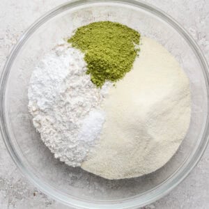 A glass bowl containing four separate dry ingredients: white flour, powdered sugar, green matcha powder, and yellowish almond flour.