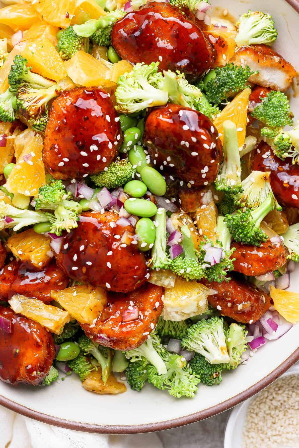 A bowl of glazed chicken with broccoli, orange slices, edamame, chopped red onion, and sesame seeds.
