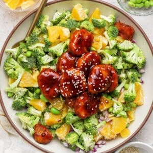A bowl of salad with broccoli, orange segments, edamame, red onion, and glazed plant-based meatballs topped with sesame seeds.