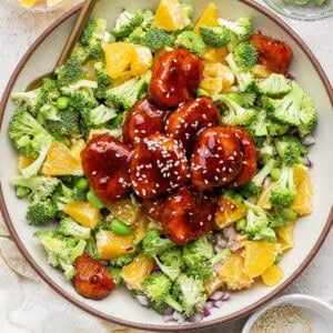 A bowl filled with broccoli, orange pieces, edamame, red onion, and glazed meatballs topped with sesame seeds, with small bowls of orange slices, edamame, and sesame seeds nearby.