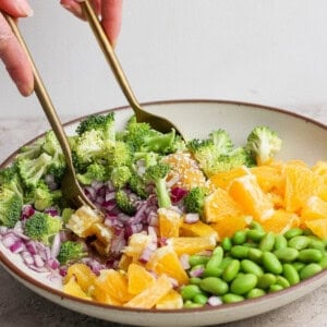 A bowl with chopped broccoli, red onion, orange segments, and edamame being mixed with two gold utensils by a person’s hands.