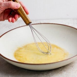 A hand holds a whisk, stirring a yellow liquid mixture in a shallow, white bowl on a light countertop.