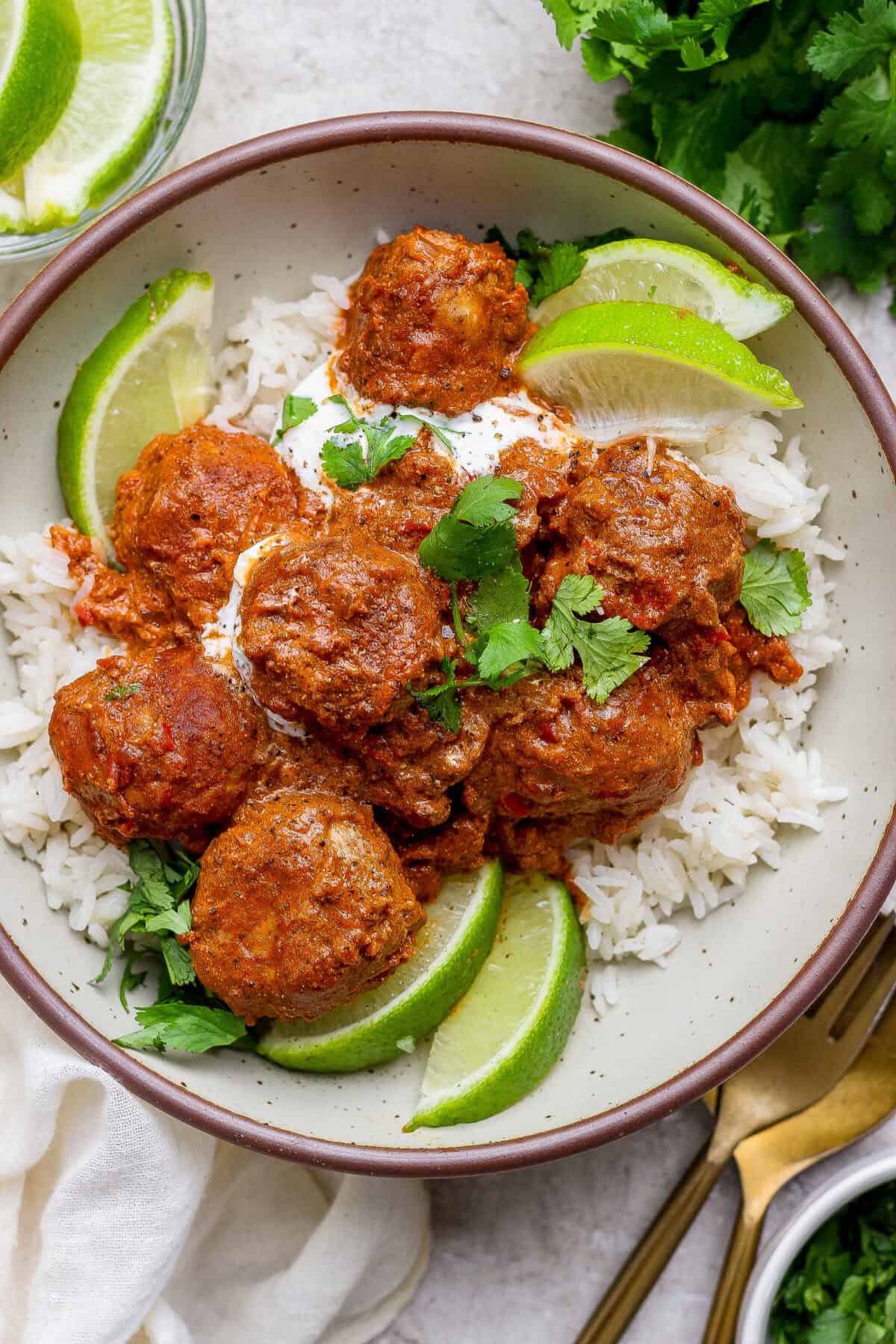 A bowl of white rice topped with butter chicken meatballs in a red sauce, garnished with lime wedges, cilantro, and a dollop of white sauce.
