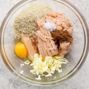 A glass bowl containing ground chicken, an egg, breadcrumbs, shredded cheese, salt, and spices—perfect for making flavorful butter chicken meatballs—sits on a light-colored surface.