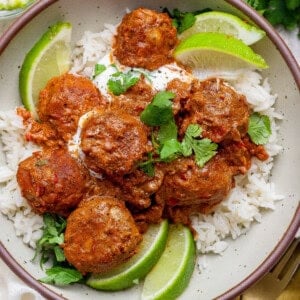 A bowl of white rice topped with butter chicken meatballs in tomato sauce, garnished with cilantro, lime wedges, and a dollop of cream.
