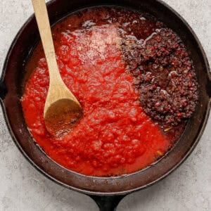 A cast iron skillet with tomato sauce, cooked ground beef, and butter chicken meatballs, stirred with a wooden spoon on a light textured surface.