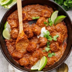 A cast iron skillet filled with butter chicken meatballs in a rich red sauce, garnished with lime wedges and cilantro, with a wooden spoon resting inside. Fresh cilantro and lime are on the side.
