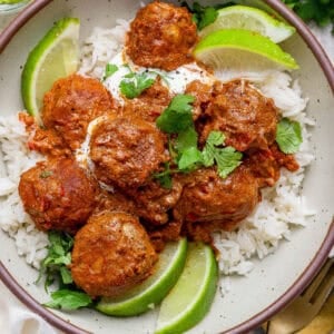 A bowl of white rice topped with butter chicken meatballs in a red sauce, garnished with lime wedges, cilantro, and a dollop of white sauce.