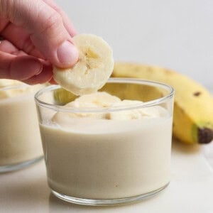 A hand placing a banana slice on top of a glass filled with banana pudding, with a whole banana in the background.