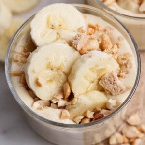 A glass bowl filled with banana pudding, topped with banana slices, chopped nuts, and crumbled cookies.