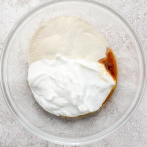 A glass bowl containing white yogurt, light brown sugar, and a pale powder on a light gray textured surface.