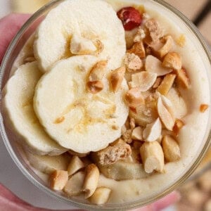 A hand holds a bowl of creamy dessert topped with banana slices and chopped peanuts.