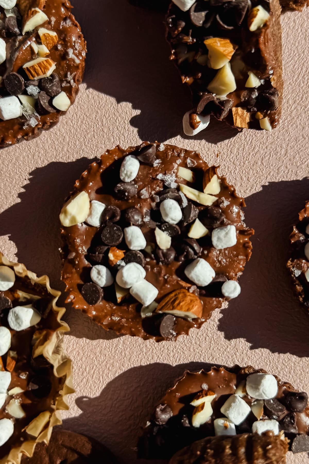 Chocolate cupcakes topped with mini marshmallows, chocolate chips, and chopped nuts, arranged on a light textured surface.