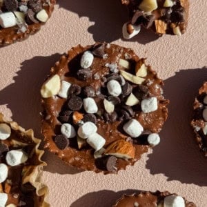 Close-up of a chocolate dessert cup topped with mini marshmallows, chocolate chips, and chopped nuts on a pink textured surface.