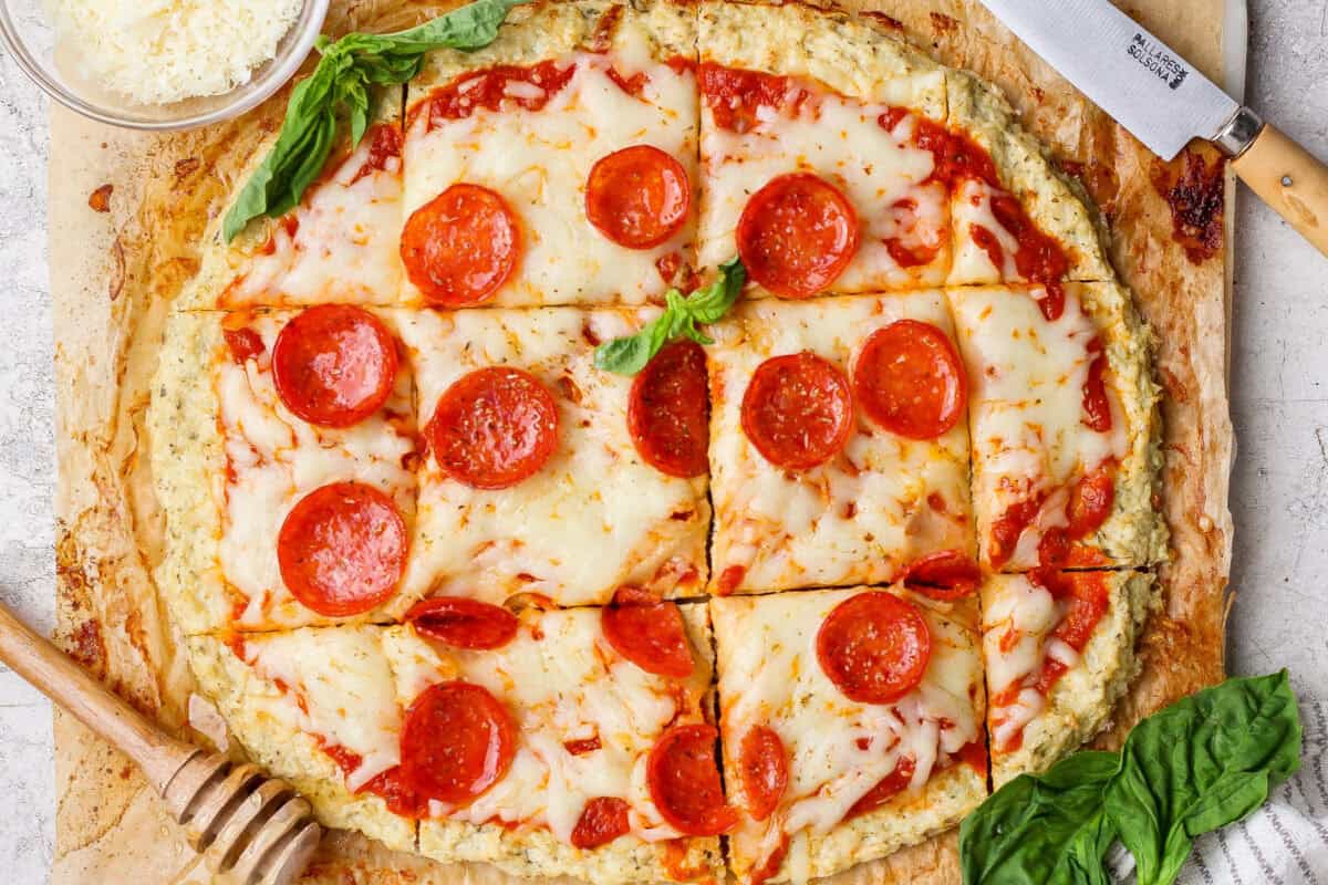 A round chicken crust pizza with melted cheese and pepperoni, cut into square slices and garnished with basil, sits next to a knife and a small bowl of grated cheese.