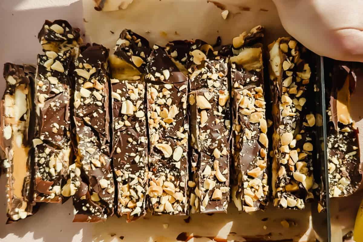 Sliced chocolate bars topped with chopped nuts are arranged on parchment paper, with a hand holding a knife to cut one of the bars.