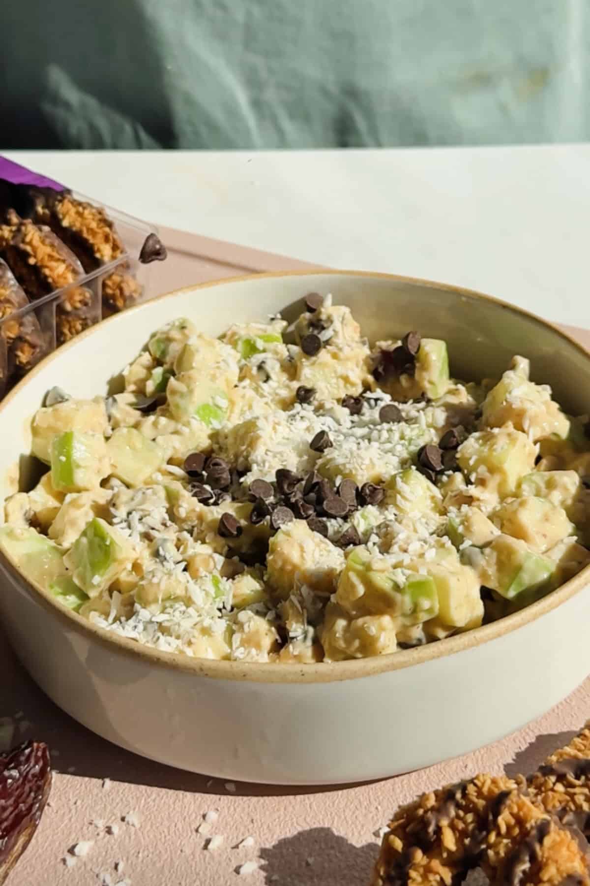 A bowl of apple salad with diced apples, shredded coconut, and chocolate chips on top, surrounded by granola bars and cookies.