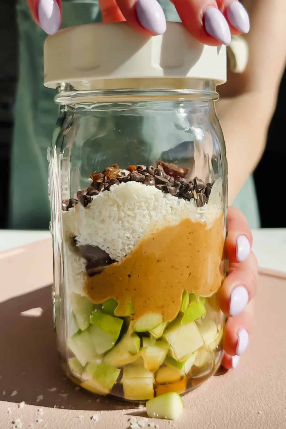 A hand holds a glass jar filled with chopped green apple, peanut butter, shredded coconut, chocolate chips, and cacao nibs, viewed from the side, with a lid being placed on top.