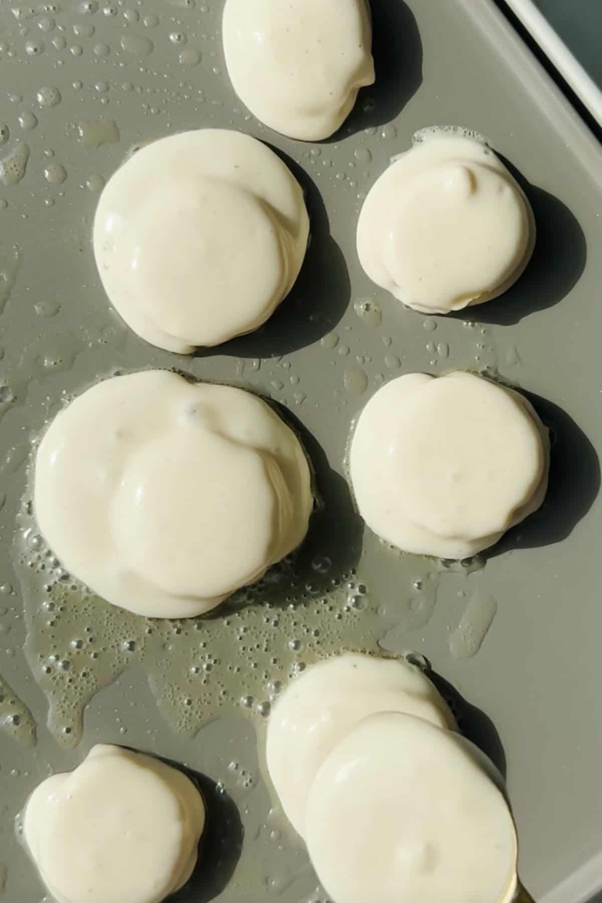 Six round dollops of pancake batter cooking on a griddle with some oil visible around the batter.