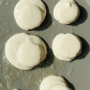 Six round dollops of pancake batter cooking on a griddle with some oil visible around the batter.