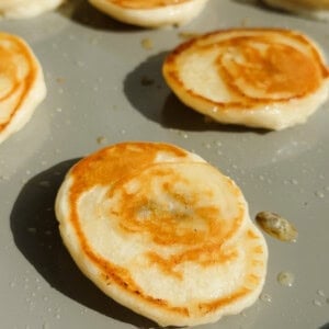 Small pancakes cooking on a lightly oiled griddle, with golden-brown surfaces and some batter droplets visible around them.