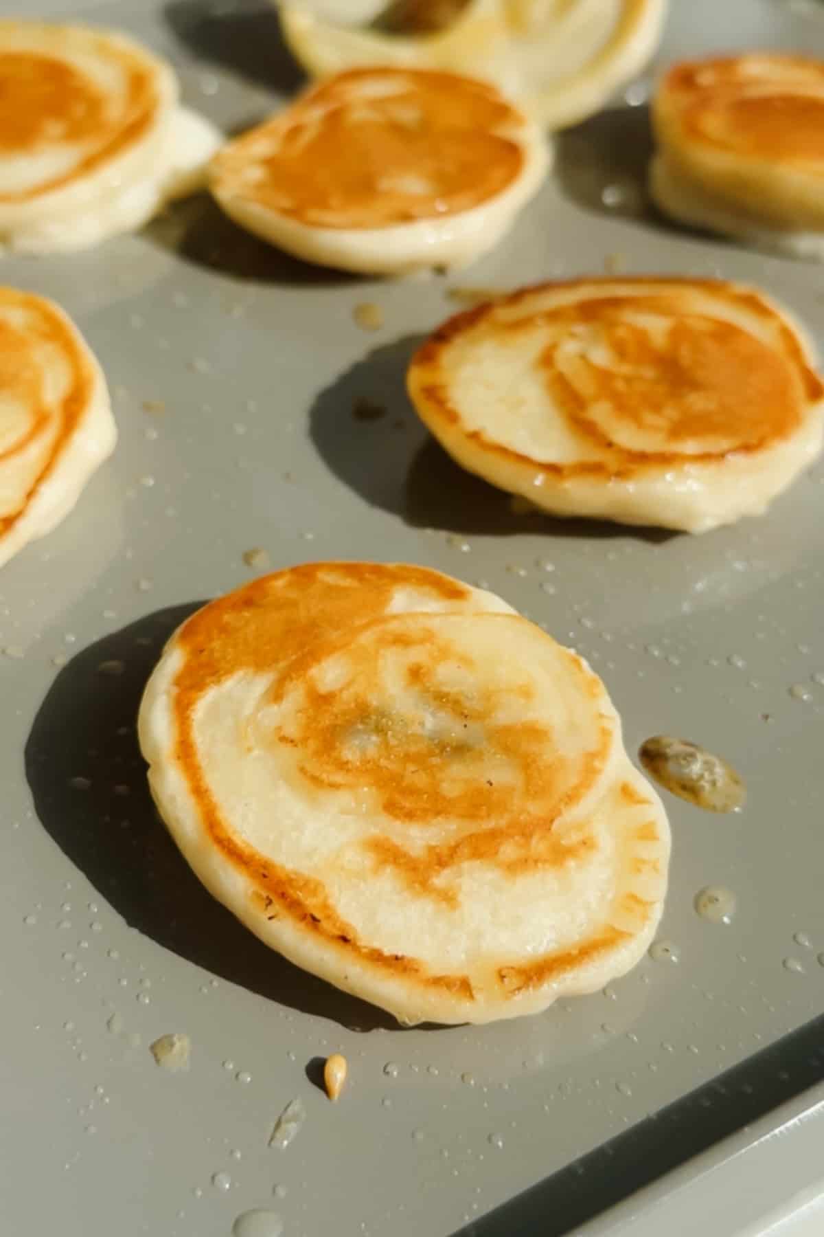 Small pancakes cooking on a lightly oiled griddle, with golden-brown surfaces and some batter droplets visible around them.