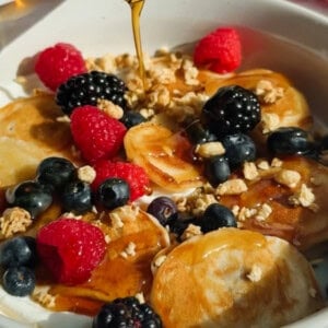A bowl of mini pancakes topped with raspberries, blackberries, blueberries, granola, and syrup being poured over them.