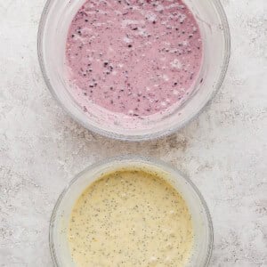 Two glass bowls on a light surface, one with purple berry-speckled batter and one with yellow batter containing small black seeds.