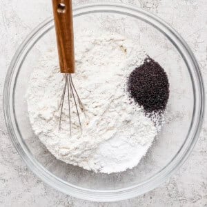 A glass bowl containing flour, baking powder, and poppy seeds with a metal whisk on a light gray surface.