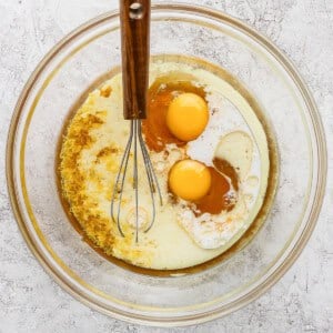 A glass mixing bowl contains two eggs, milk, oil, lemon zest, and vanilla extract with a whisk resting inside.