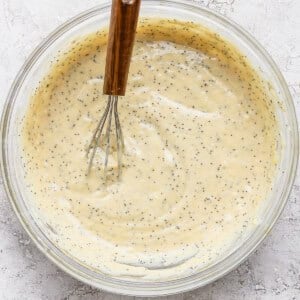 A glass bowl filled with poppy seed batter is shown with a metal whisk with a wooden handle resting inside.