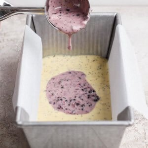 A metal scoop pours purple berry batter over yellow batter in a parchment-lined loaf pan on a light surface.