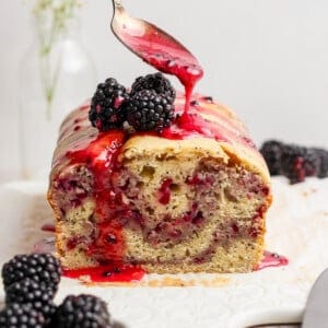 A hand drizzles berry sauce over a sliced blackberry loaf cake topped with fresh blackberries on a white cutting board.