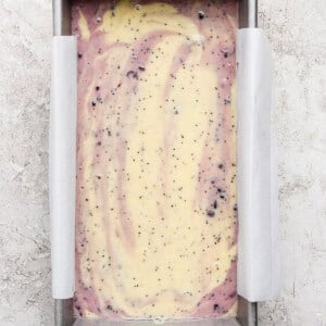 Rectangular loaf pan lined with parchment paper, filled with swirled yellow and purple cake batter containing small dark specks, on a light textured surface.