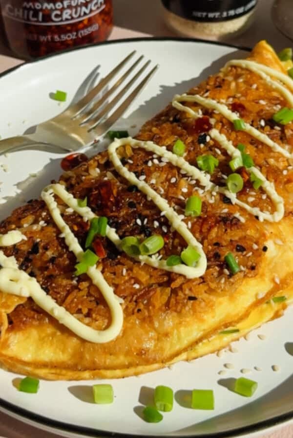 A golden omelette filled with fried rice, topped with mayonnaise, chopped green onions, sesame seeds, and chili flakes on a white plate with a fork.