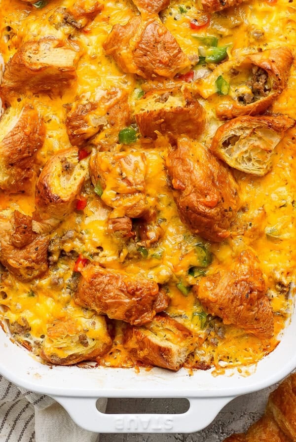 A casserole dish filled with a baked croissant breakfast casserole topped with melted cheese and diced vegetables on a textured surface. A white striped cloth is partially visible.