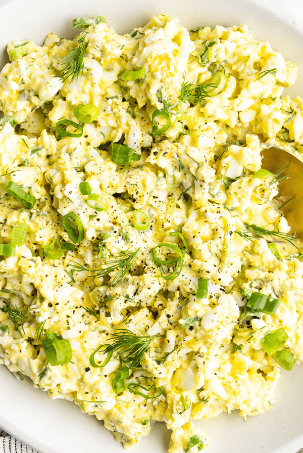 A bowl of egg salad with chopped green onions and dill, accompanied by a gold spoon.