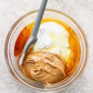 A glass bowl containing peanut butter, yogurt, honey, and an egg with a gray spatula resting inside.