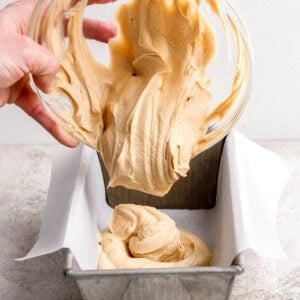 A person pours thick, light brown batter from a glass bowl into a parchment-lined metal loaf pan on a light-colored surface.