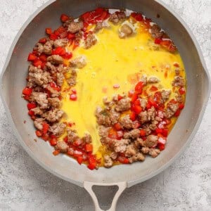 A skillet containing cooked sausage, diced red bell peppers, and beaten eggs beginning to cook.