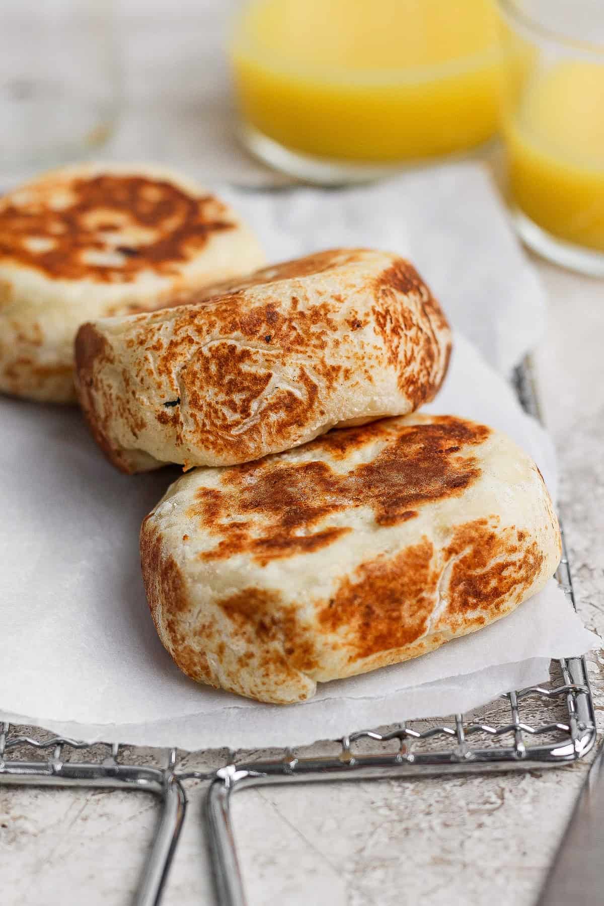 Three browned, thick English muffins rest on parchment paper atop a cooling rack, with blurred glasses of orange juice in the background.