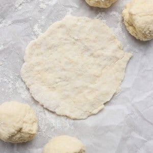 Four dough balls and one flattened round of dough sit on a sheet of parchment paper dusted with flour.