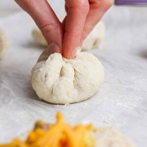 A hand pinches the top of a ball of dough to seal it closed; fillings and shredded cheese are visible on a piece of dough in the foreground.