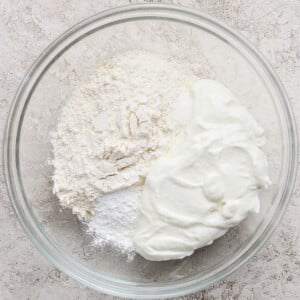 A glass bowl containing flour, baking powder, salt, and plain yogurt on a textured light surface.
