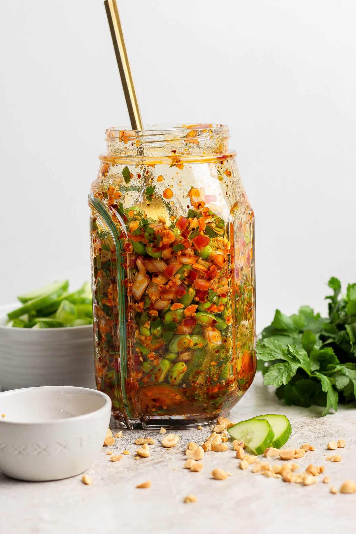 A glass jar filled with a colorful chopped vegetable salad, topped with sauce and a spoon, sits on a countertop with herbs, peanuts, and cucumber slices nearby.